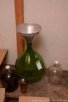 Green glass jug with metal funnel, dark jug, clear jug with cap, and amber bottle grouped on floor near wall.