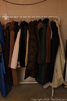 Photo of approximately 12 ladies coats hanging on a rack in neutral colored room. Varying colors and materials visible including wool, faux leather, nylon. Some coats have fur collars or hoods.