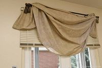 Beige patterned curtain draped loosely over window blinds, curtain rod included. Shows fabric texture and pattern.