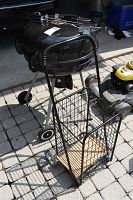 Photo of black charcoal kettle grill on tripod stand with wheels, wheeled metal buggy in front with wooden base, and barbecue utensils placed on grill lid.