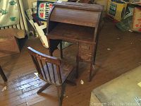 Photo showing vintage wooden children's roll top desk and matching wooden swivel chair on wooden floor with some boxes in background