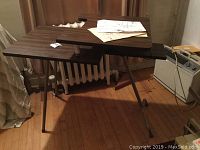 Side angle of vintage two tier sewing machine folding table showing dark wood grain surface and metal foldable legs with assembly instructions on top.