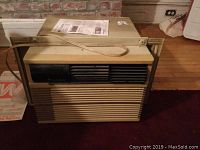 Front view of the air conditioner showing beige body, black vent grille, and power cord on top.