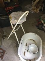 Image showing white enamel baby tub with rust and wear, including a small matching enamel bowl inside. Background includes a white stool with cloth cover (not included).