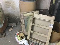Photo showing two wooden vintage rustic wall cabinet doors with chipped paint and wear, one with glass panel, stacked with other items on the floor