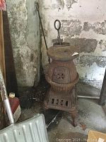 Full view of an antique cast iron pot belly stove with flat top, four legs, and an operating lever visible at the top, sitting on a basement floor near wall pipes and assorted tools.