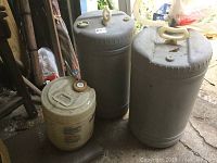 Three kerosene tanks, two large grayish-white 15-gallon tanks and one smaller beige 5-gallon tank. Tanks show signs of use with some dirt and wear.