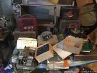 Overall lot showing several radios, parts, and stacks of journals on a cluttered surface in a basement or storage area.