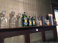 Shelf display of multiple collectible liquor, liqueur, and champagne bottles, including clear cut-glass decanter bottles on the left, several green and clear international liqueur bottles, and novelty coffee thermoses on the right.