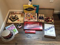 Photo showing arranged collection of board games and puzzles on wooden floor, including Scrabble, Sequence, Yahtzee, Billiard Ball Rummy, Mickey Mouse puzzle, Word Search book, Dominoes, and marbles.