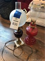 Photo showing two vintage oil lamps, one with white marble base and etched frosted globe, the other with red textured glass base and clear chimney, placed on wooden floor.