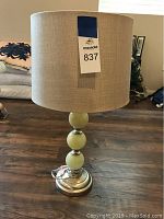 Front view of the table lamp showing beige linen-look barrel shade and three greenish polished orb base with metal accents and round metal base on a wood floor.