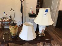 Two Asian inspired table lamps on a wooden table, one with a dark base and botanical theme, the other with an Asian figurative design and floral patterns. Both have white wide fabric lampshades.