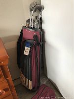 Full view of burgundy Max Fli golf bag filled with Pal Joey golf clubs and a folded umbrella lying in front