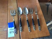 Five pieces of vintage silver-toned serveware including two serving spoons and three serving forks with black handles and ornate silver detailing on the ends laid on wooden surface.