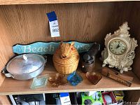 Shelf display showing aluminum serving dish, wicker honey pot, beach house sign, glass dishes, vintage pepper grinder, vintage cigarette dispenser, and an ornate decorative clock.