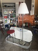 Floor lamp with brushed nickel finish, white pleated shade, and attached glass table with wrought iron chair featuring red upholstered seat positioned beside it.