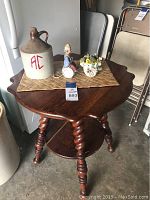 Full view of carved wooden table with carved, twisted legs, split lower shelf, and items on top including a jug, clown nightlight, ceramic floral arrangement, and woven placemat.