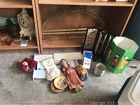 Photo showing collection of dolls, decorative plates, three-panel screen, ceramic planter, and figurine head on concrete floor in front of wooden shelves.