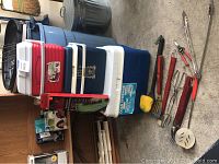 Three coolers stacked including a red and blue small can cooler and a large blue and white 34-quart Kooler ice chest cooler alongside various BBQ utensils laid out on concrete floor.
