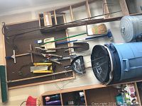 Wide shot of various garden tools hung on pegboard including a broom, pry bars, shovels, a dustpan, and a blue plastic trashcan and small metal trashcan beneath them
