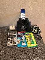 Overview of lot showing black Swift binoculars, vintage camera, Sharp printing calculator with attached paper roll, and two tubes of household cement glue in packaging.