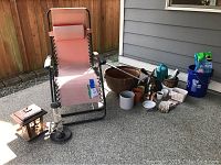 Photo of outdoor recliner lounger with pink fabric and black metal frame, adjacent to various garden items including pots, soil bag, bird feeders, and yard decorations arranged on concrete floor against wooden fence and house siding.