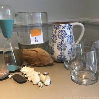 Photo of the entire lot showing ceramic pitcher, wine glasses, hourglass, wooden fish, shells, stones, and the hurricane vase.
