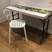 Folding table with black metal legs and white top, white metal chair, and floral fabric runner on table visible from front and side angle showing details.