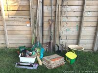 Photo showing assorted garden tools including shovels, rakes, hoe, ceramic planter, lawn bags, and miscellaneous items next to a wooden fence on grass.