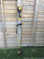 Full length view of the Dewalt 20V Max XR rechargeable trimmer leaning against a wooden fence with safety guard at the cutting end, adjustable handle, and battery pack on top.