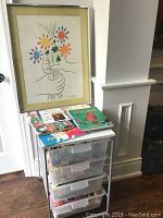 Photo of framed Picasso print, stack of books on top of IKEA storage unit with plastic drawers containing art supplies