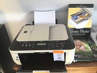 Canon MX320 printer in white and black, with assorted photo and inkjet paper packs to the right side, including a glossy photo paper pack.