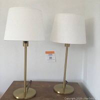 Pair of brass finish IKEA table lamps with white fabric lampshades shown side by side on a wooden surface against a white wall.