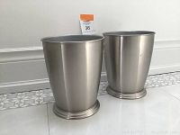 Two brushed nickel metal waste baskets side by side on tiled floor against white baseboard, showing shape and finish
