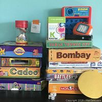 Photo of stacked table and board games including Cranium, Cadoo, Monopoly NHL Edition, Battleship, Boggle electronic game, Perfection game plastic unit, educational electronic kits, and a table tennis set.