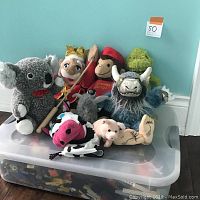 Photo showing various puppets and stuffed toys arranged on top of a clear plastic bin; puppets include king, Curious George, koala, green plush, blue furry creature, cow, pig