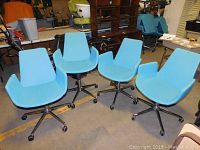 Four turquoise office chairs viewed from a frontal angle showing unique seat shape, chrome bases, caster wheels, and padded upholstery.