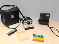 View of two Polaroid instant cameras; one white OneStep camera with rainbow stripe, one black Sun 600 LMS camera. Accompanying accessories: black carrying bag, flash bar, Time-Zero Supercolor film, Polaroid booklet.
