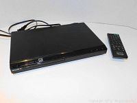 Front angled view of black Sony DVD player with power cord and remote control on white surface.