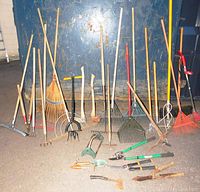 Wide shot of various yard cleanup tools including rakes, hoes, axes, and hand tools arranged against a blue wall on a concrete floor.