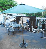 Green outdoor patio umbrella fully opened with metal pole and decorative base, outdoor setting with other patio furniture and equipment in background.