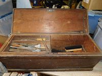 Open antique wooden tool chest showing various vintage hand tools inside, including chisels and brushes.