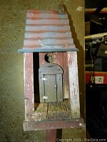 Front view of wooden bird house showing small door with metal hinges and latch, red corrugated metal roof, and wooden pole.