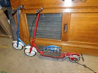 Side view of two vintage metal Kick 'N Go scooters, one red and one blue, showing footboards, wheels, and chain drive areas.