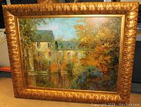 Front view of the oil painting showing the house and trees near water with golden ornate frame.
