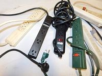 Photo showing six assorted surge protectors with plugs and cords in various colors and styles.