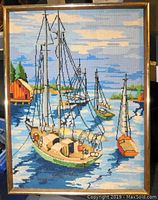 Framed needlepoint artwork showing sailboats docked on water with surrounding landscape.