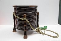 Front side view of round Japanese storage box with dark lacquer and bronze accents on feet, showing attached green braided rope.