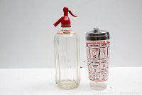 Crystal Springs soda siphon bottle with red spout and multi-color cocktail shaker side by side on white background